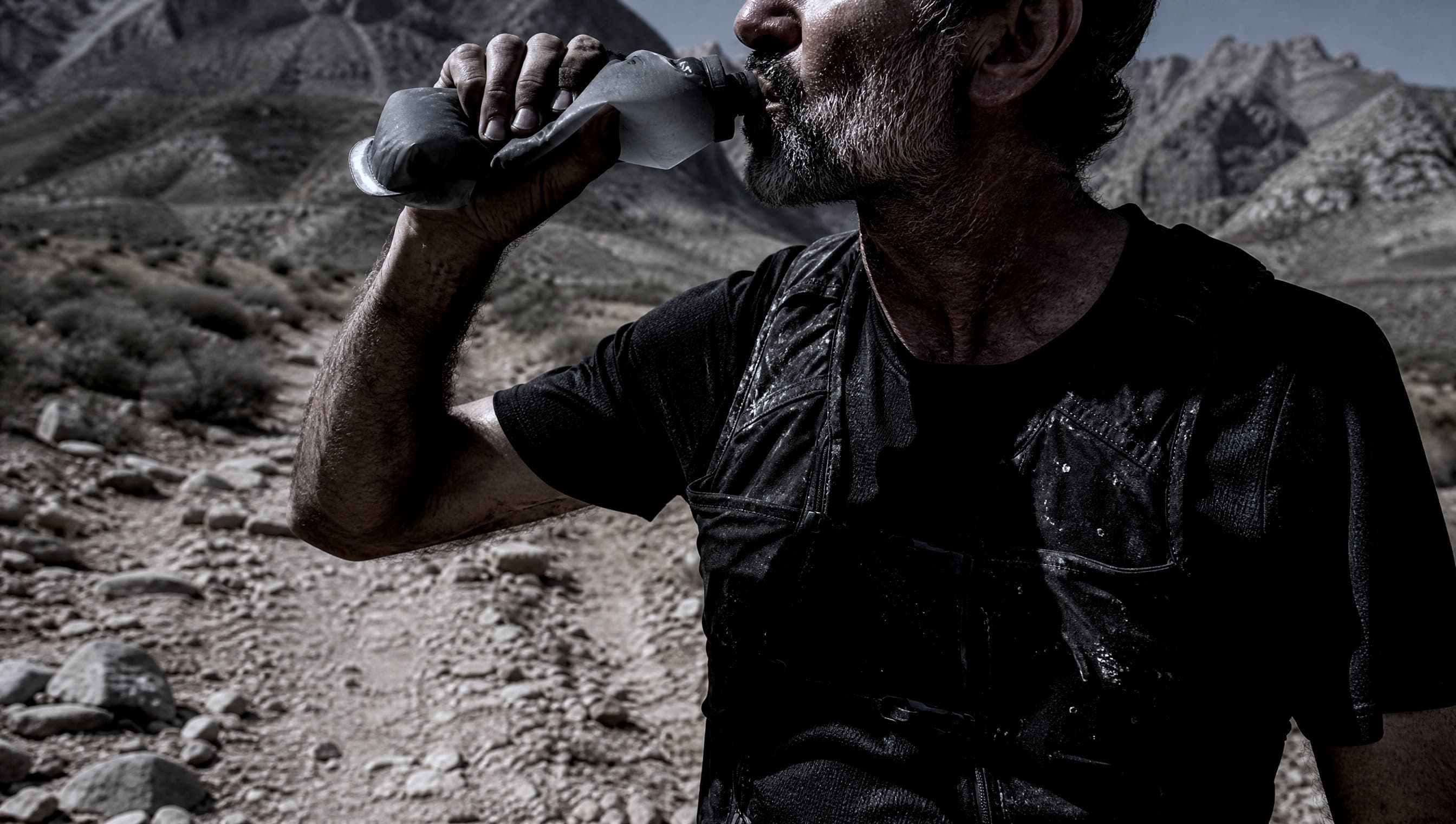 A masters-age trail runner drinking from a soft flask on a dusty stony track in harsh midday light