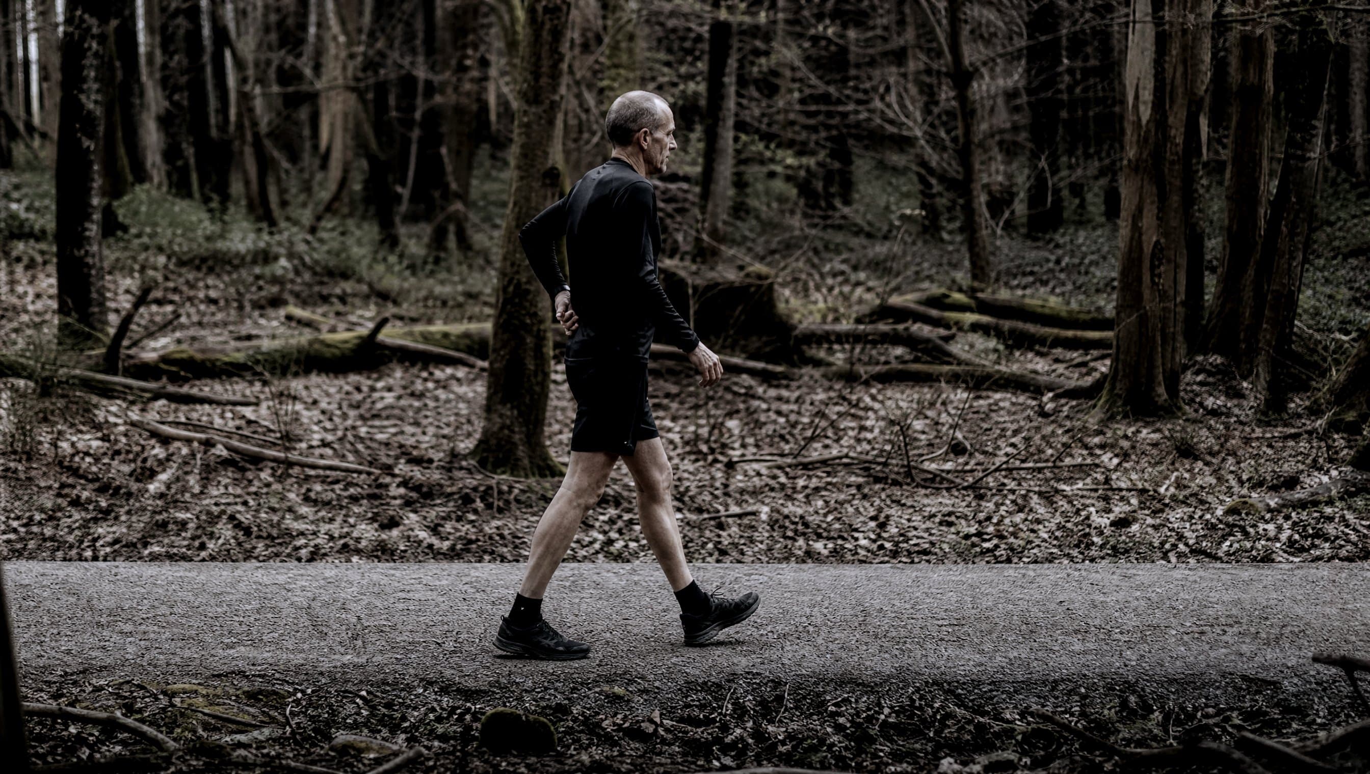 A masters-age runner walking rather than running along a flat gravel trail through bare woodland
