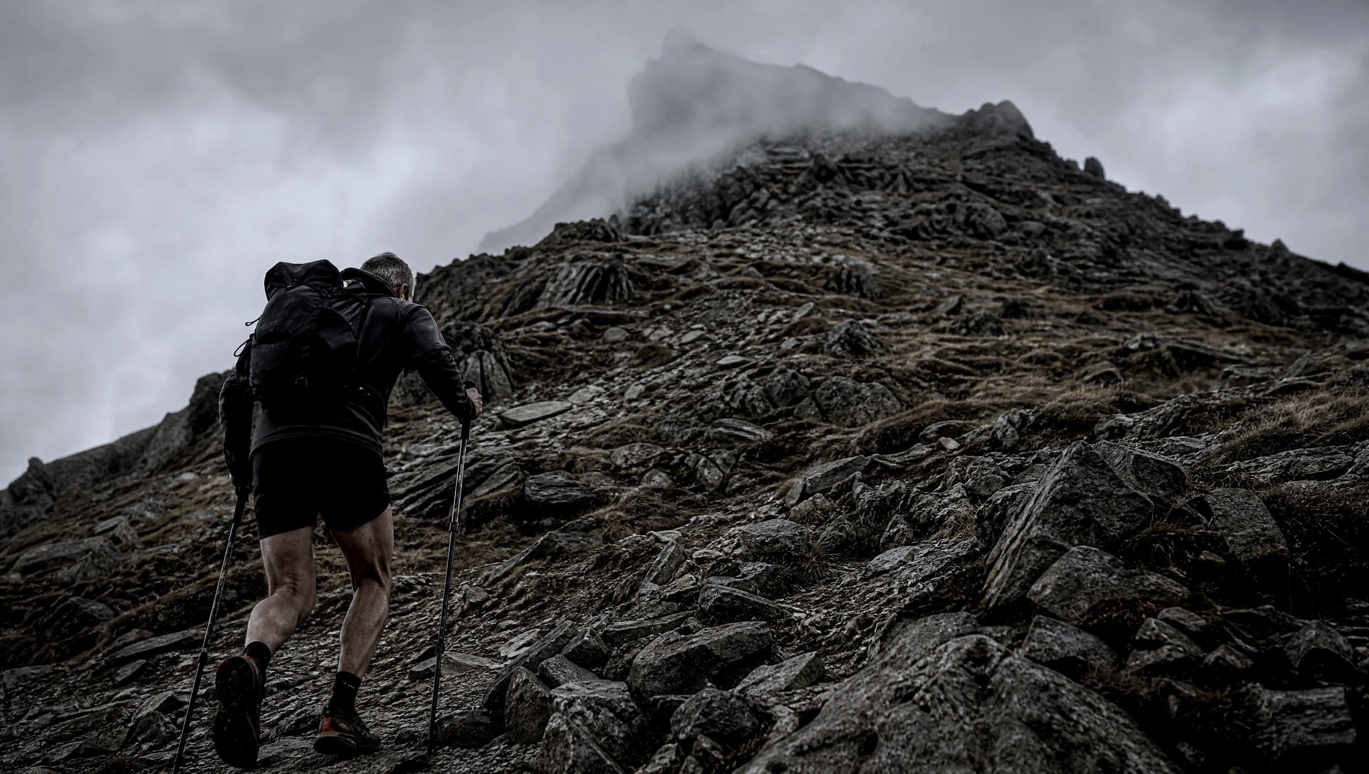 A masters-age runner power hiking a steep rocky climb with a loaded pack and poles, seen from behind