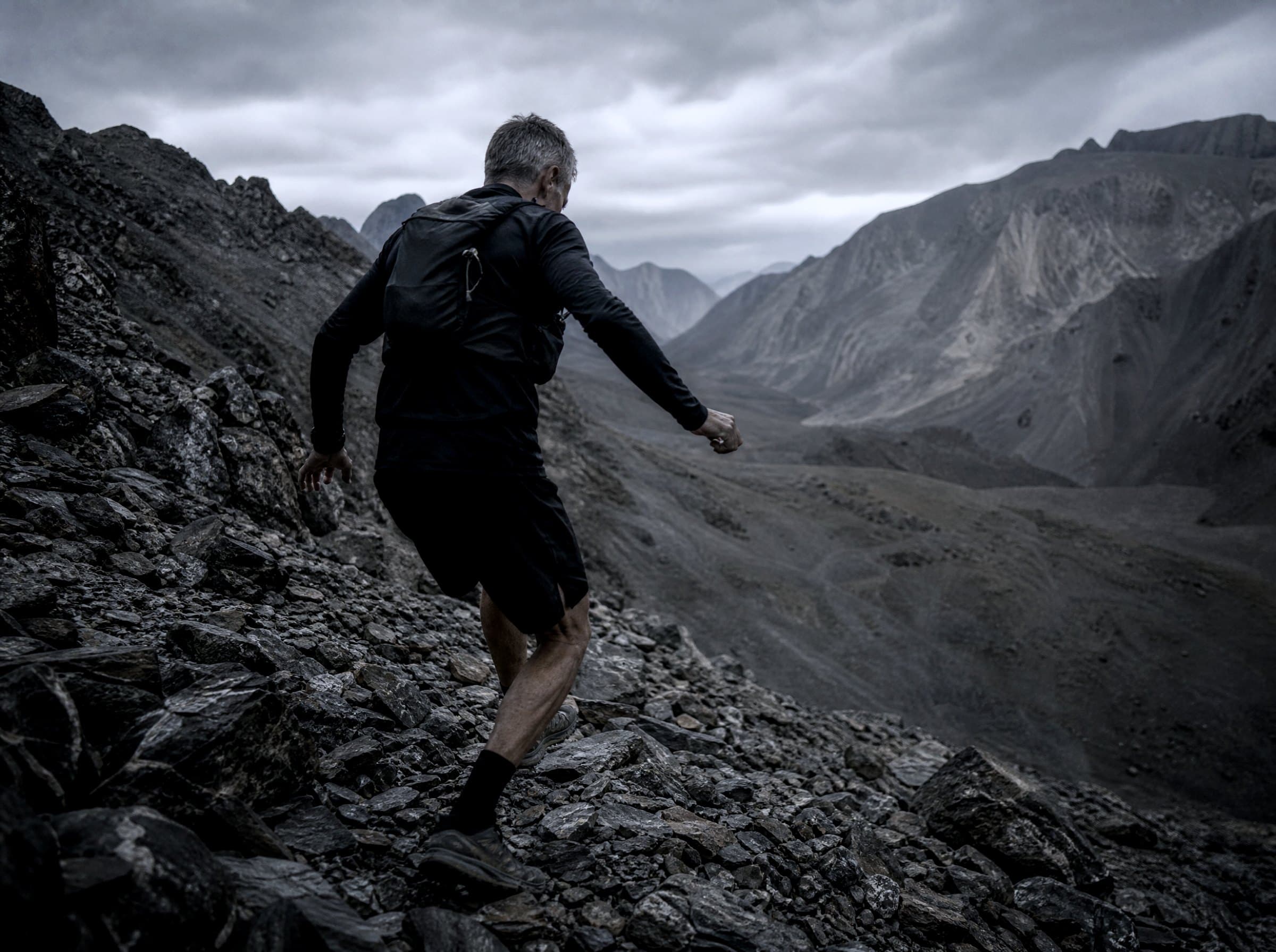 A runner moving through grey alpine scree under heavy cloud, mountains falling away behind