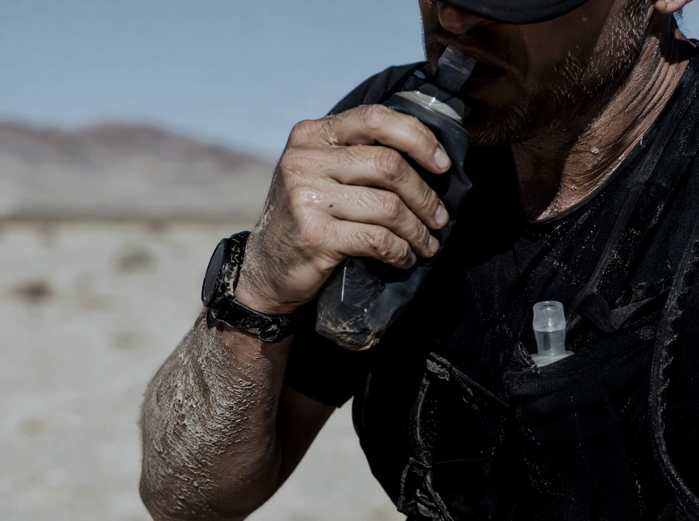 An ultra runner in desert heat squeezing a soft flask to drink, salt crust on the forearm