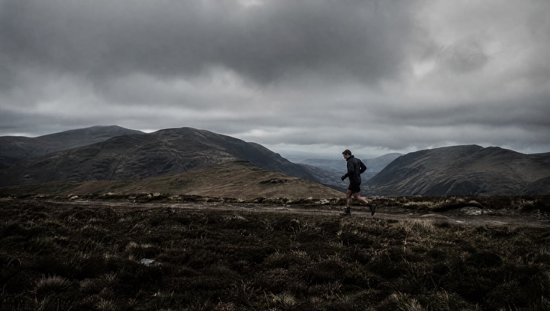 An ultra runner deep into a long run on an exposed moorland ridge under heavy skies, tired but still moving