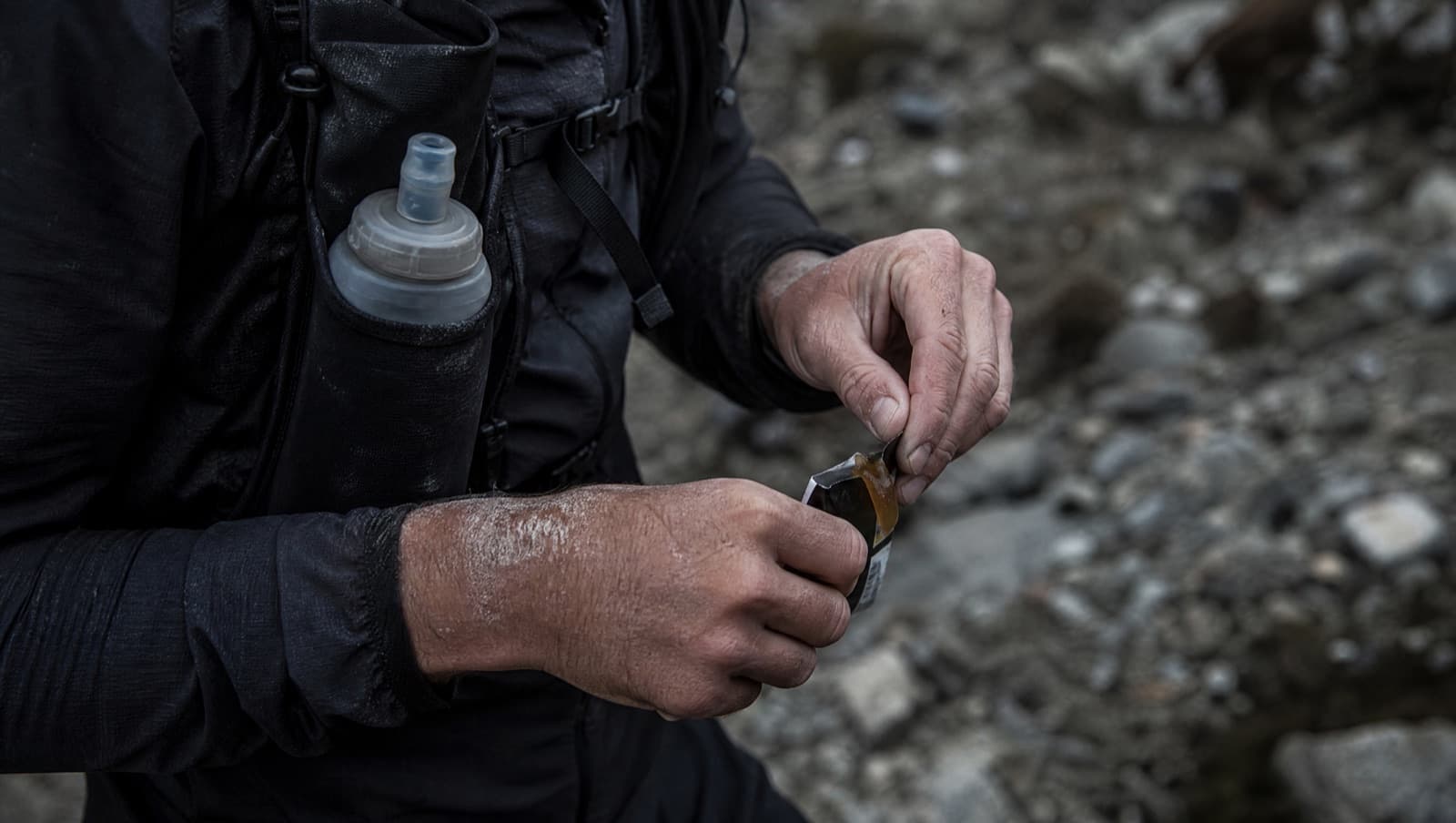 An ultra runner's hands tearing open an energy gel mid-run on a rocky trail