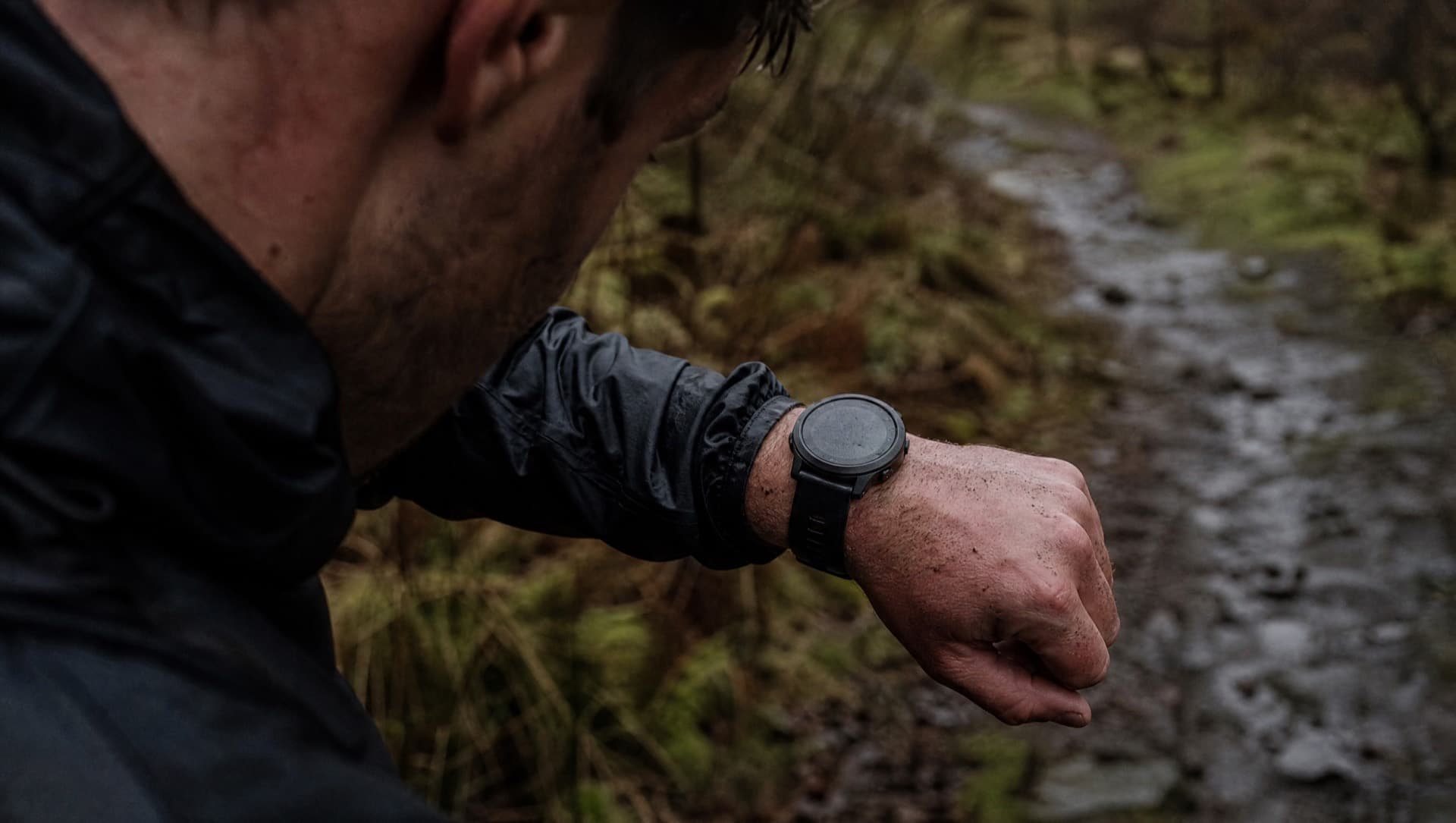 A close crop of a tired trail runner checking a plain GPS watch, mud on the forearm, wet woodland behind