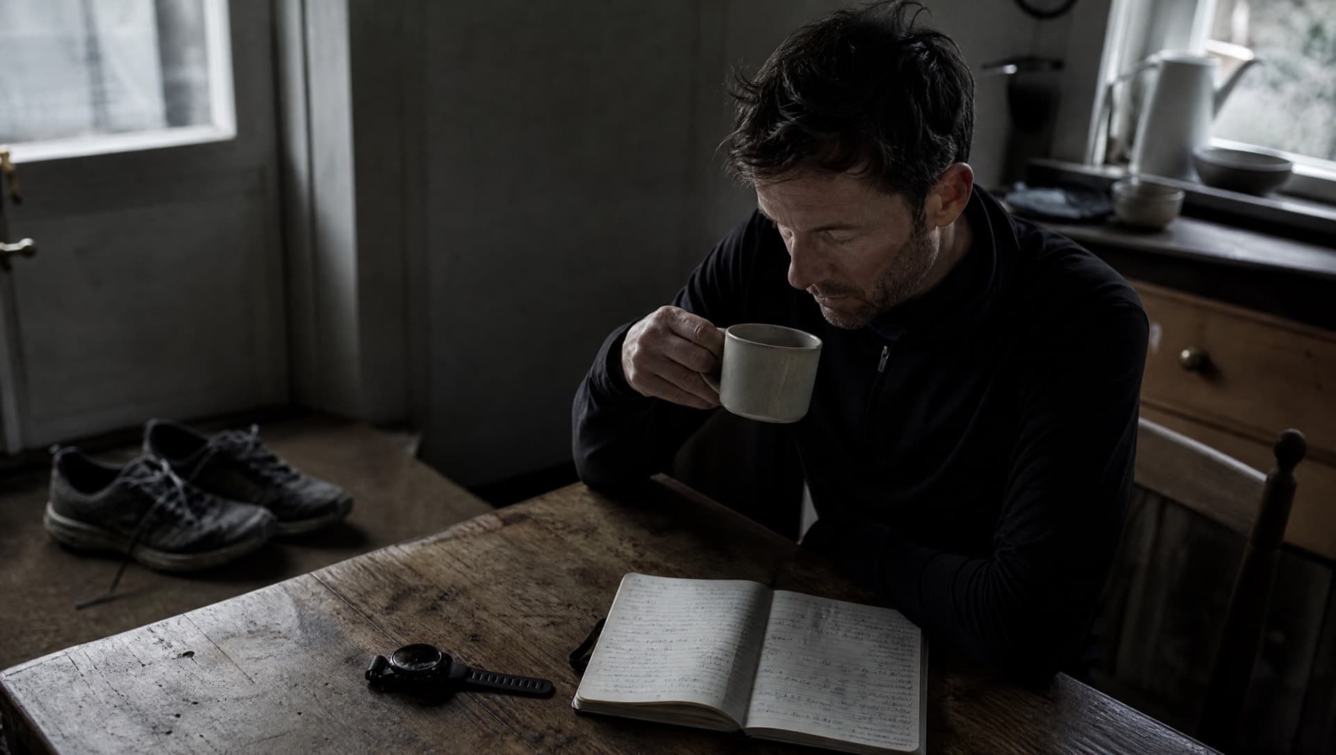 A masters runner at a kitchen table in early morning light with coffee, a paper training diary and a GPS watch