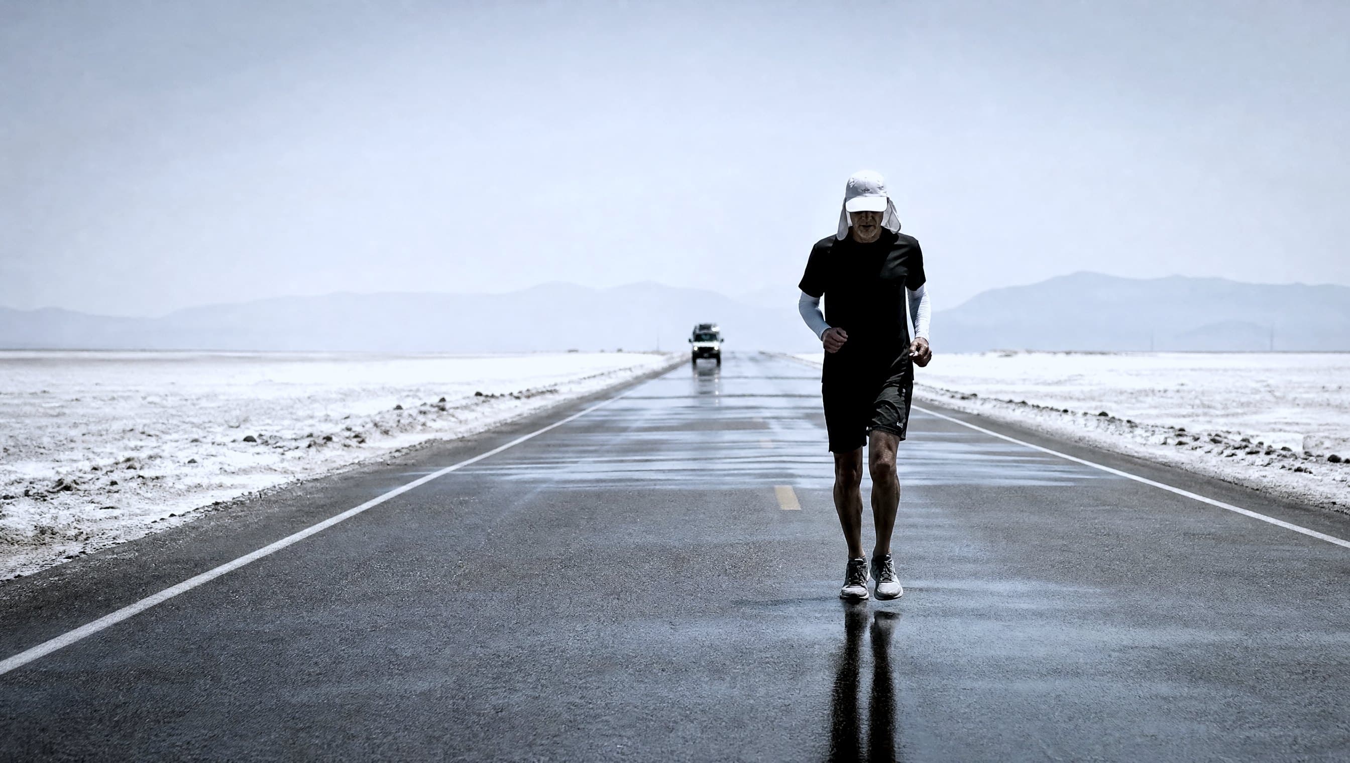 A masters-age runner in white on a straight desert highway across a salt flat in extreme heat