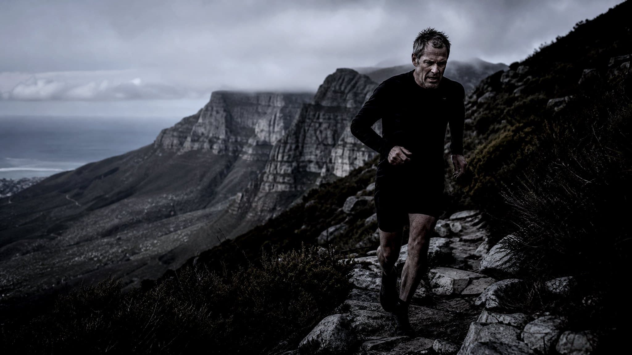 A masters-age trail runner on a rocky mountain path above Cape Town with cliffs and the sea below