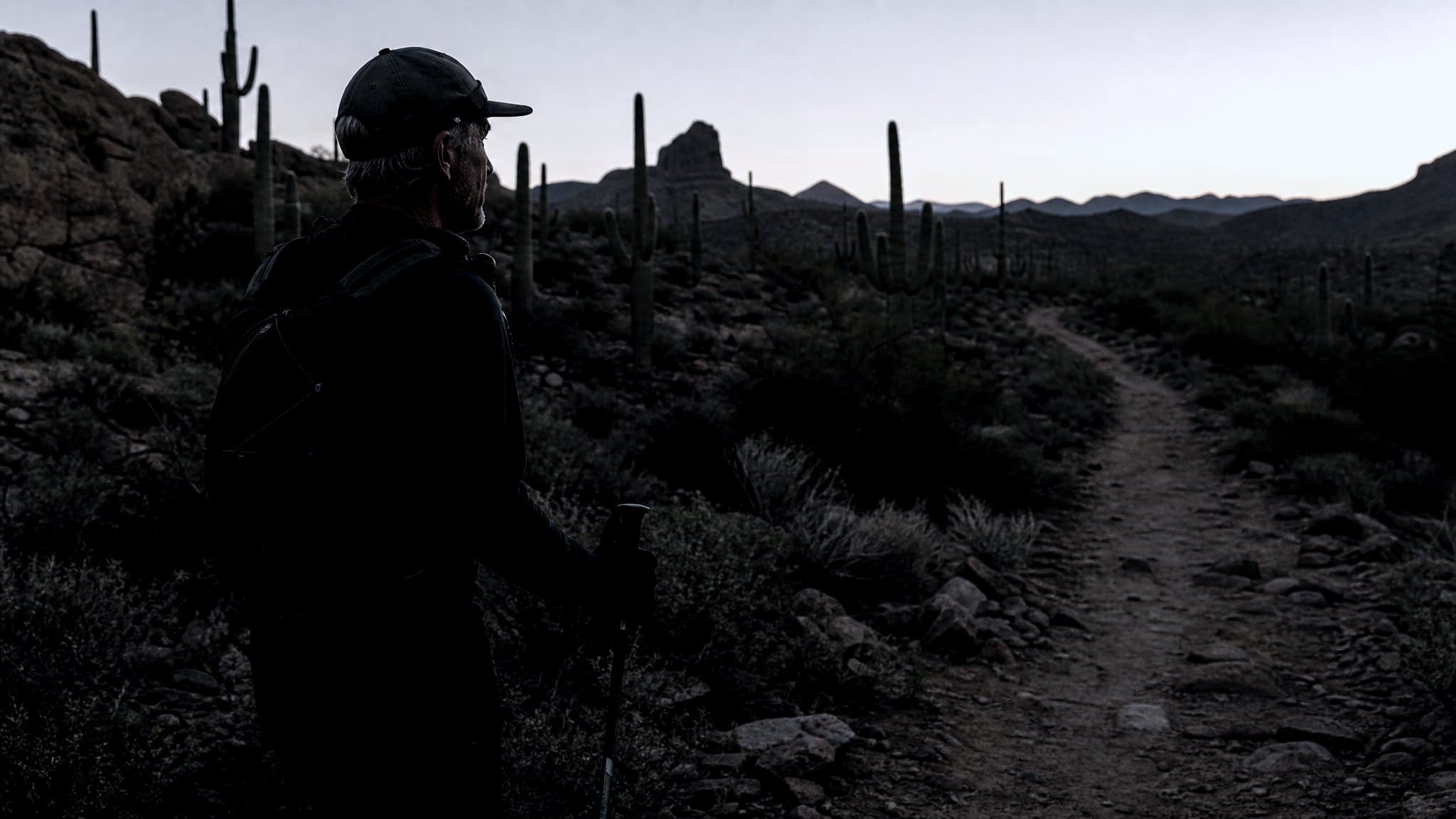 A masters-age runner walking through saguaro cactus and red rock at first light with poles and a small pack