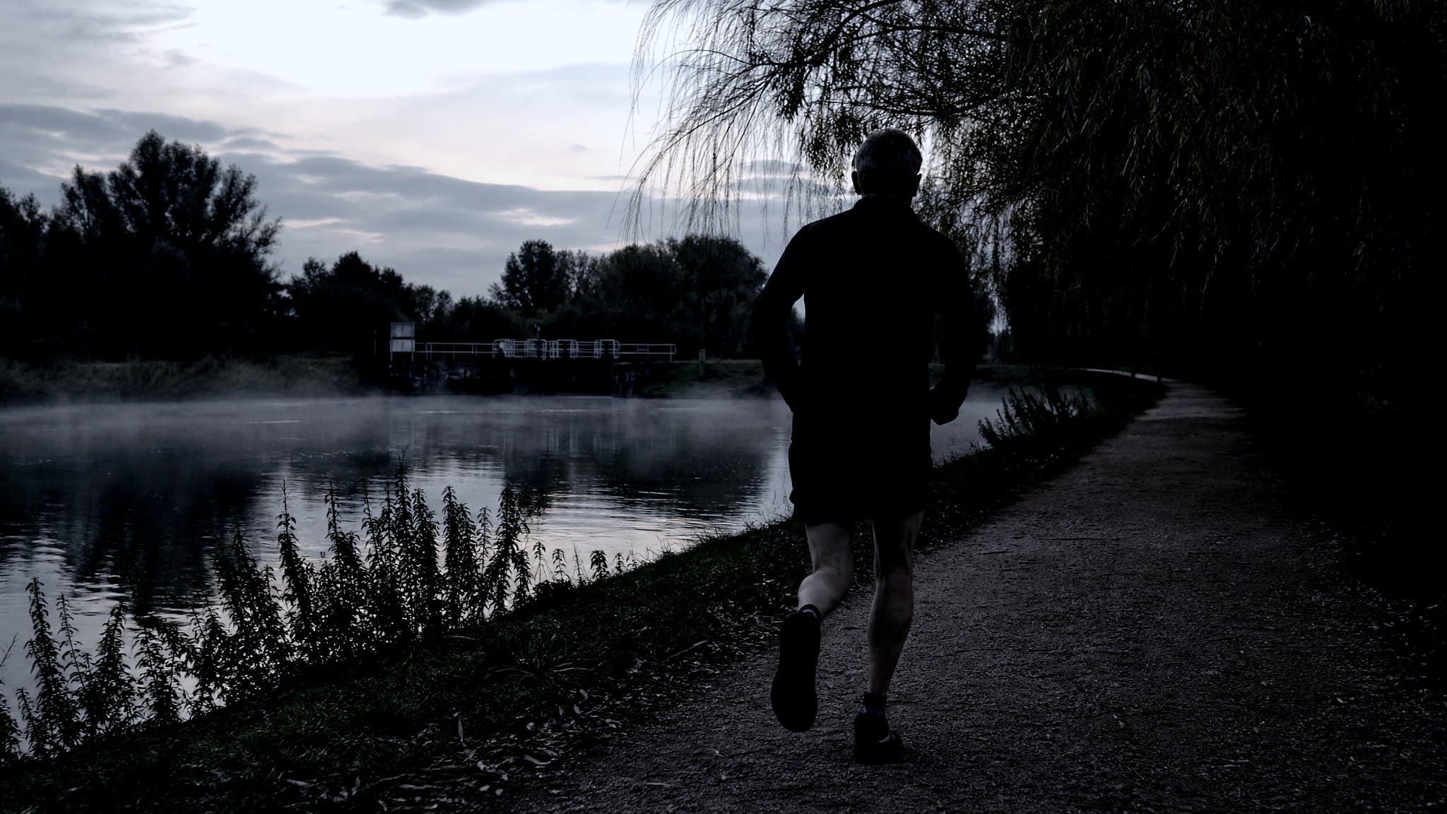 A masters-age runner on a flat gravel towpath beside the Thames at dusk with mist on the water
