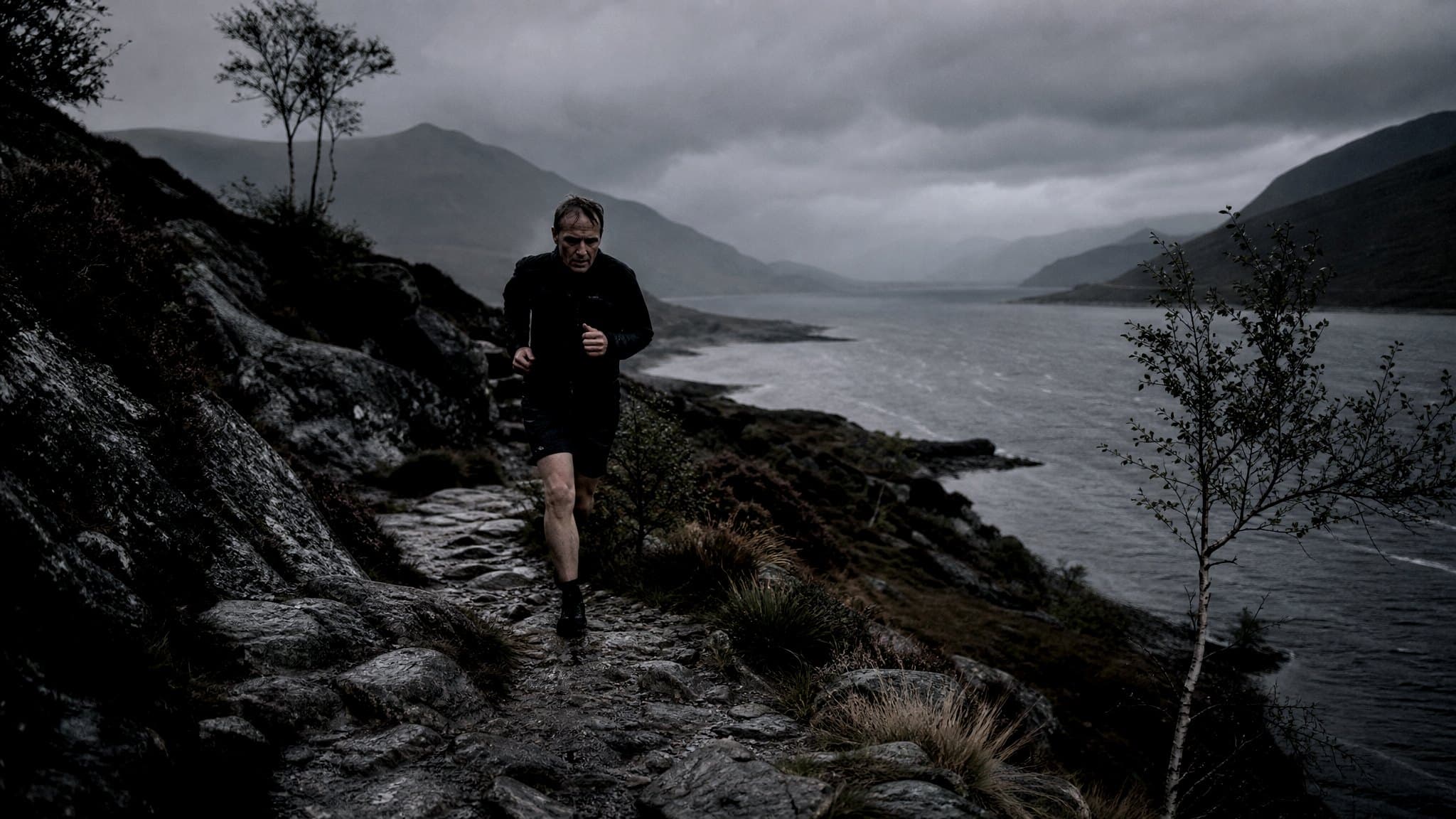 A masters-age runner on a stony path along a Scottish loch shore under low grey cloud